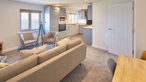 A modern kitchen with white cabinets, a dining area with a wooden table, and a living room with a beige sofa and chairs. - Host Stay Sea Watcher (Whitby)