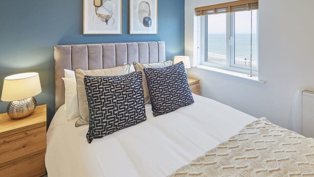 A neatly made bed with patterned pillows, a bedside lamp, and a view of the ocean through a window. - Host Stay Sea Watcher (Whitby)