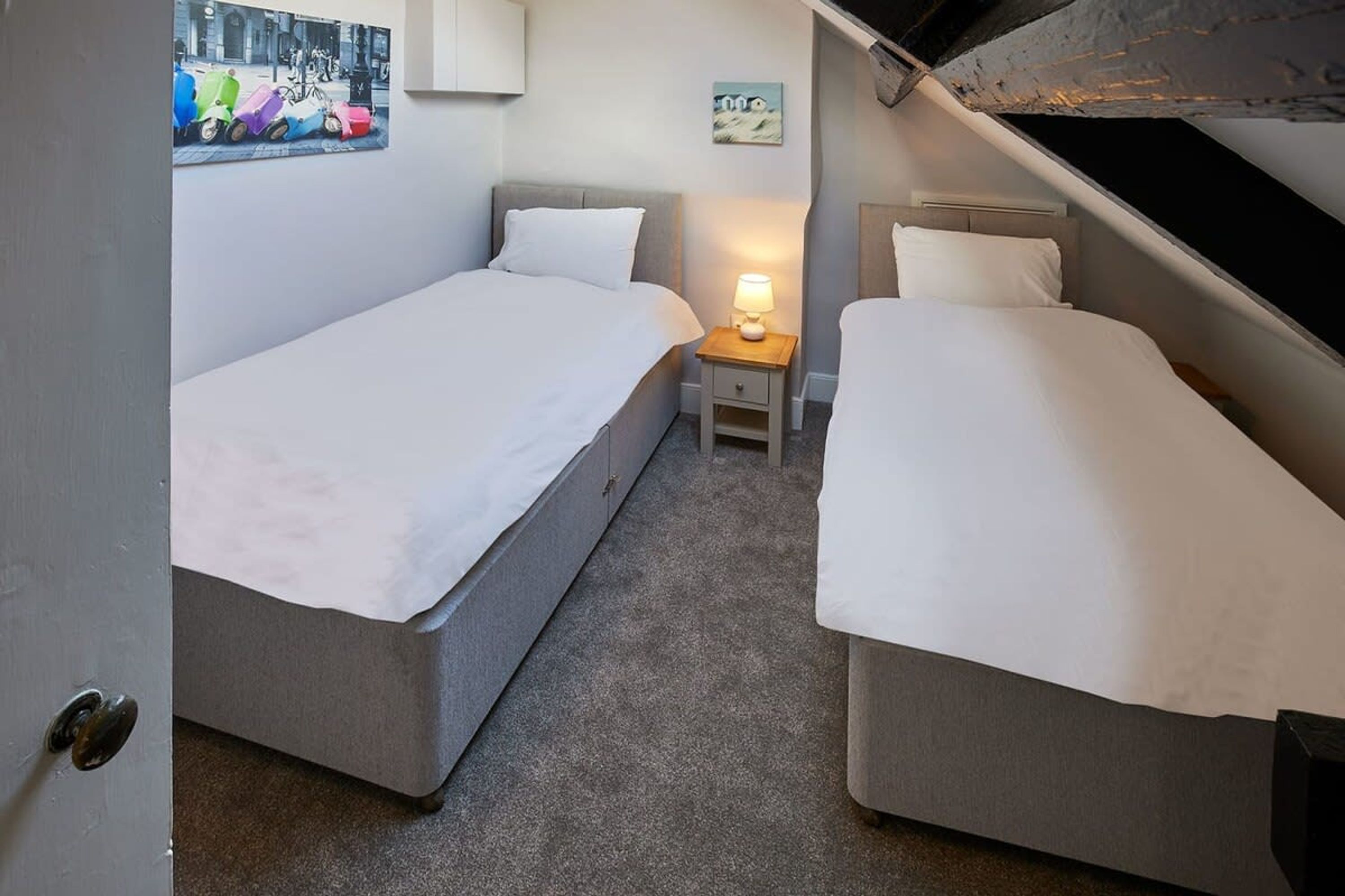Une petite chambre à coucher, de taille réduite, avec deux lits simples, une table de chevet avec une lampe et un tableau accroché au mur.