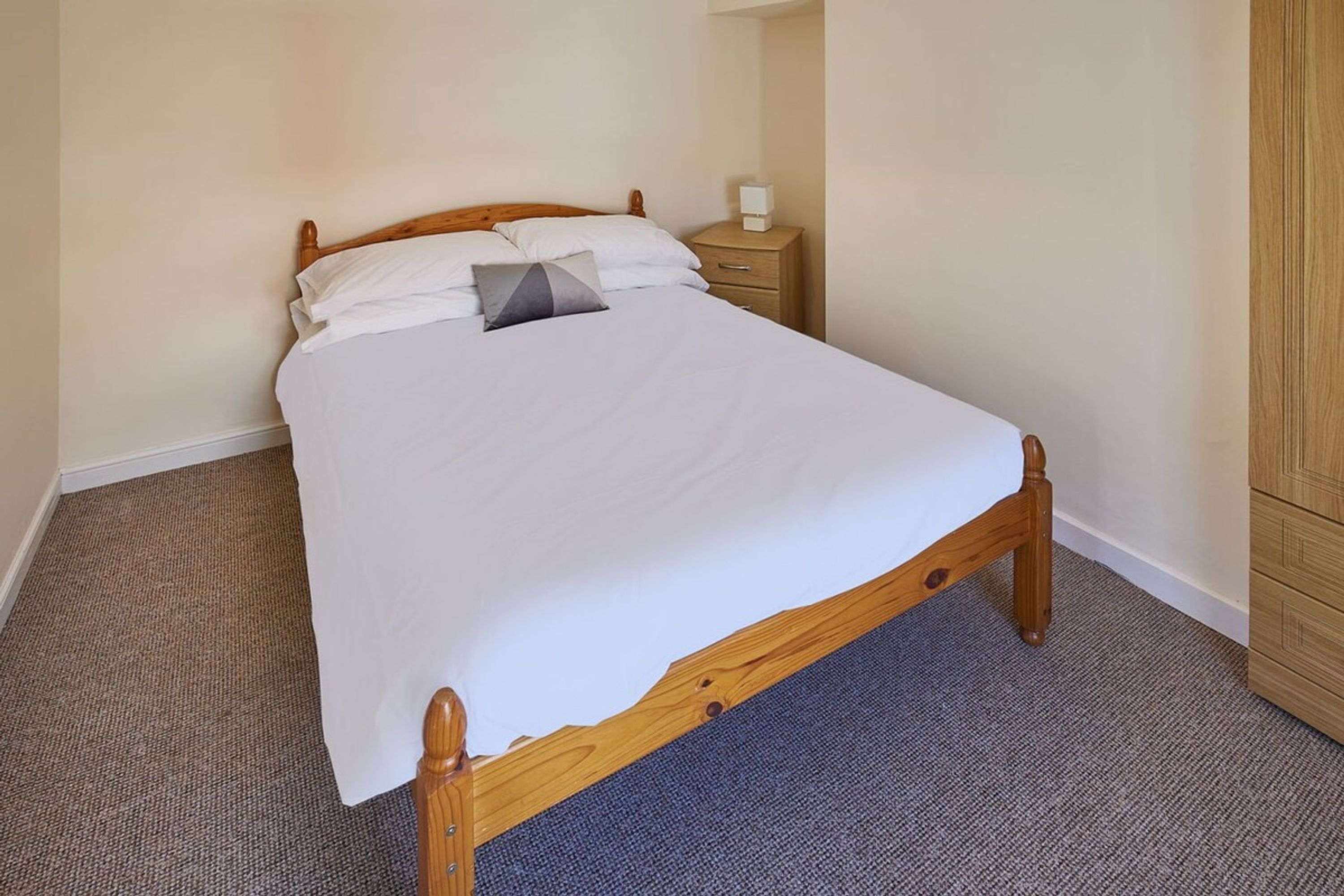 A single bed with a wooden frame and white bedding, a bedside table with a lamp, and a wardrobe.