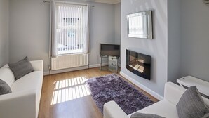 A modern living room with a fireplace, a television on a stand, and a large window with curtains. - Host Stay 1 Oswy Street (Whitby)