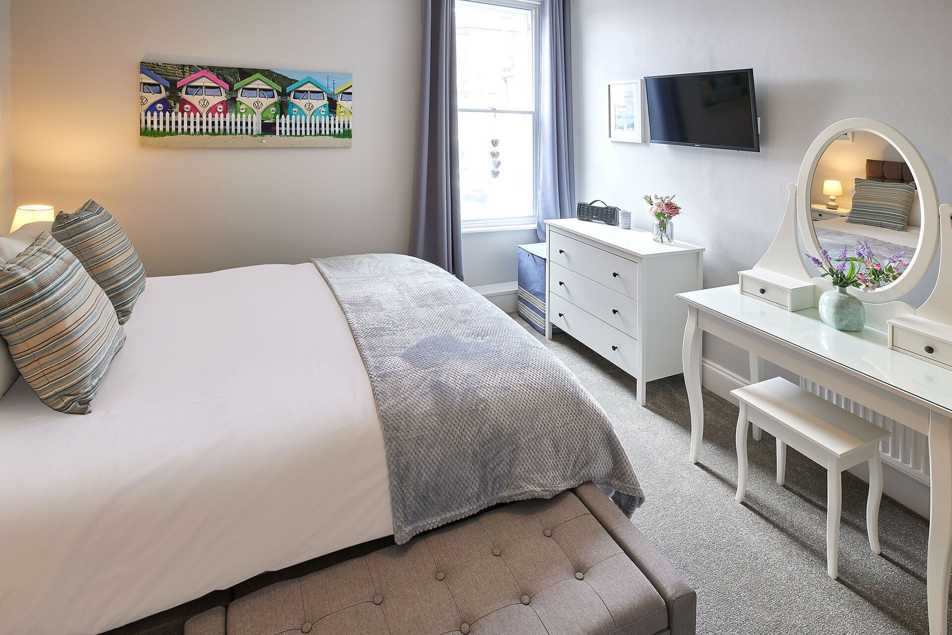 A bedroom with a bed, dresser, vanity, and a painting on the wall.