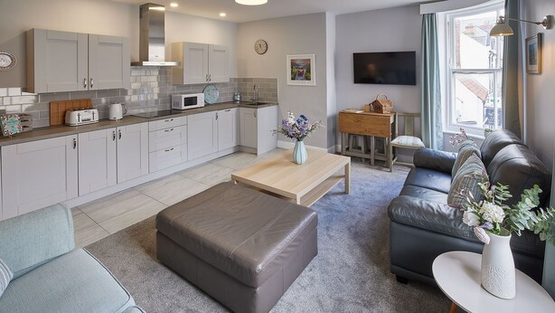 A modern kitchen with white cabinets, a microwave, and a toaster. There is a wooden coffee table with a vase of flowers. A grey sofa and a black leather couch are in the living area, which has a grey carpet. A TV is mounted on the wall, and a window with curtains is visible. - Host Stay The Runswick at Brunswick Holiday Apartments (Whitby)