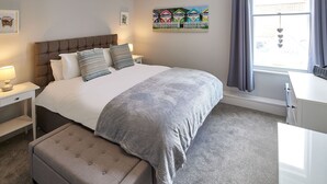 A neatly arranged bedroom with a large bed, a bedside table with a lamp, a bench, and a window with curtains. - Host Stay The Runswick at Brunswick Holiday Apartments (Whitby)