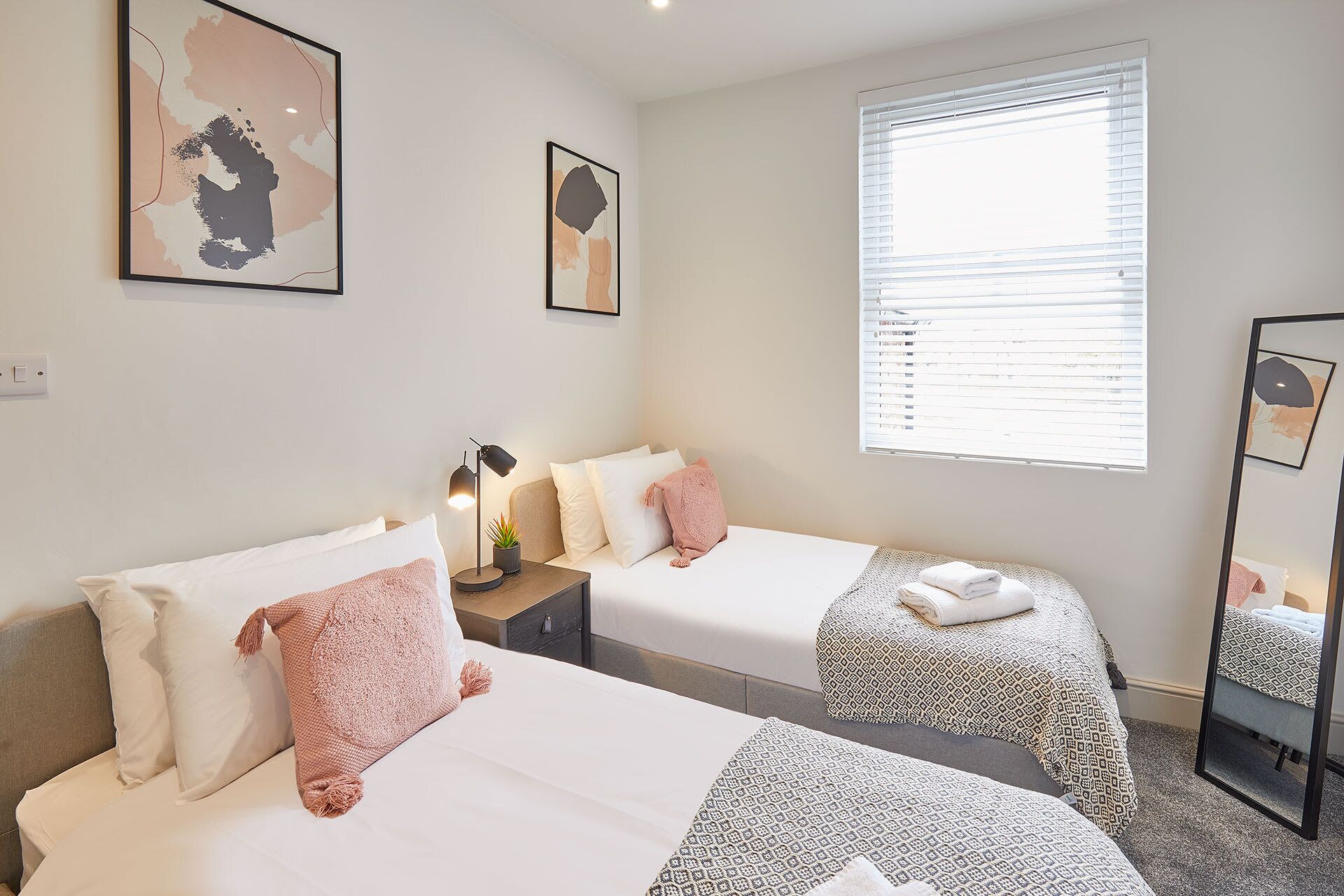 Une chambre d’hôtel avec deux lits, une grande fenêtre avec des stores, une table de chevet avec une lampe et des œuvres d’art accrochées aux murs.