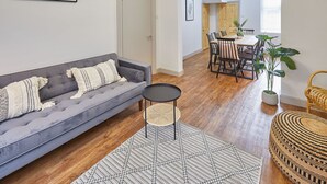 Una sala de estar con un sofá gris, una mesita redonda pequeña, un tapete tejido y un comedor con una mesa y sillas de madera.