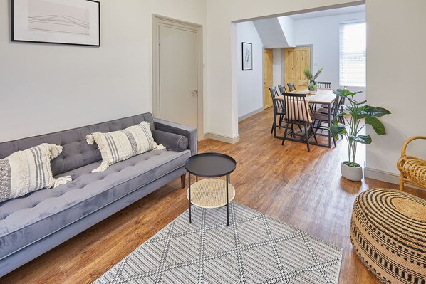 A living room with a grey sofa, a small round table, a woven rug, and a dining area with a wooden table and chairs.