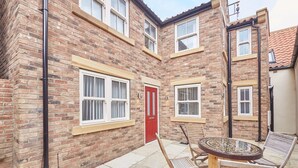 A brick building with a red door, two windows with white frames, and a round table with chairs in the courtyard. - Host Stay The Cottage Bagdale Hall (Whitby)