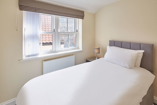 A neatly made bed with a grey headboard, a window with blinds, and a bedside lamp.