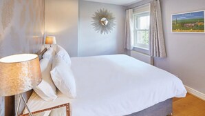 Une chambre à coucher avec un lit, une lampe, un miroir, une fenêtre avec des rideaux et un tableau accroché au mur.