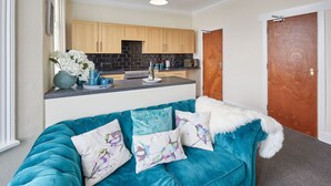 A teal sofa with decorative pillows, a kitchen area with wooden cabinets and black backsplash, and a wooden door.