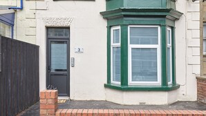 A corner of a building with a dark door, a window with green shutters, and a brick wall.