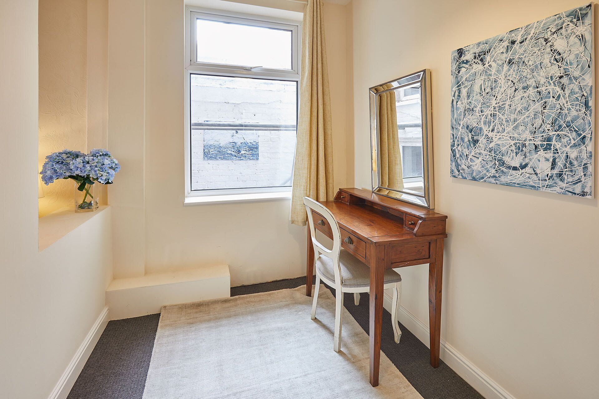 Une pièce comprenant une coiffeuse en bois, un miroir, une fenêtre avec des rideaux, un vase de fleurs et une œuvre d’art abstraite accrochée au mur.