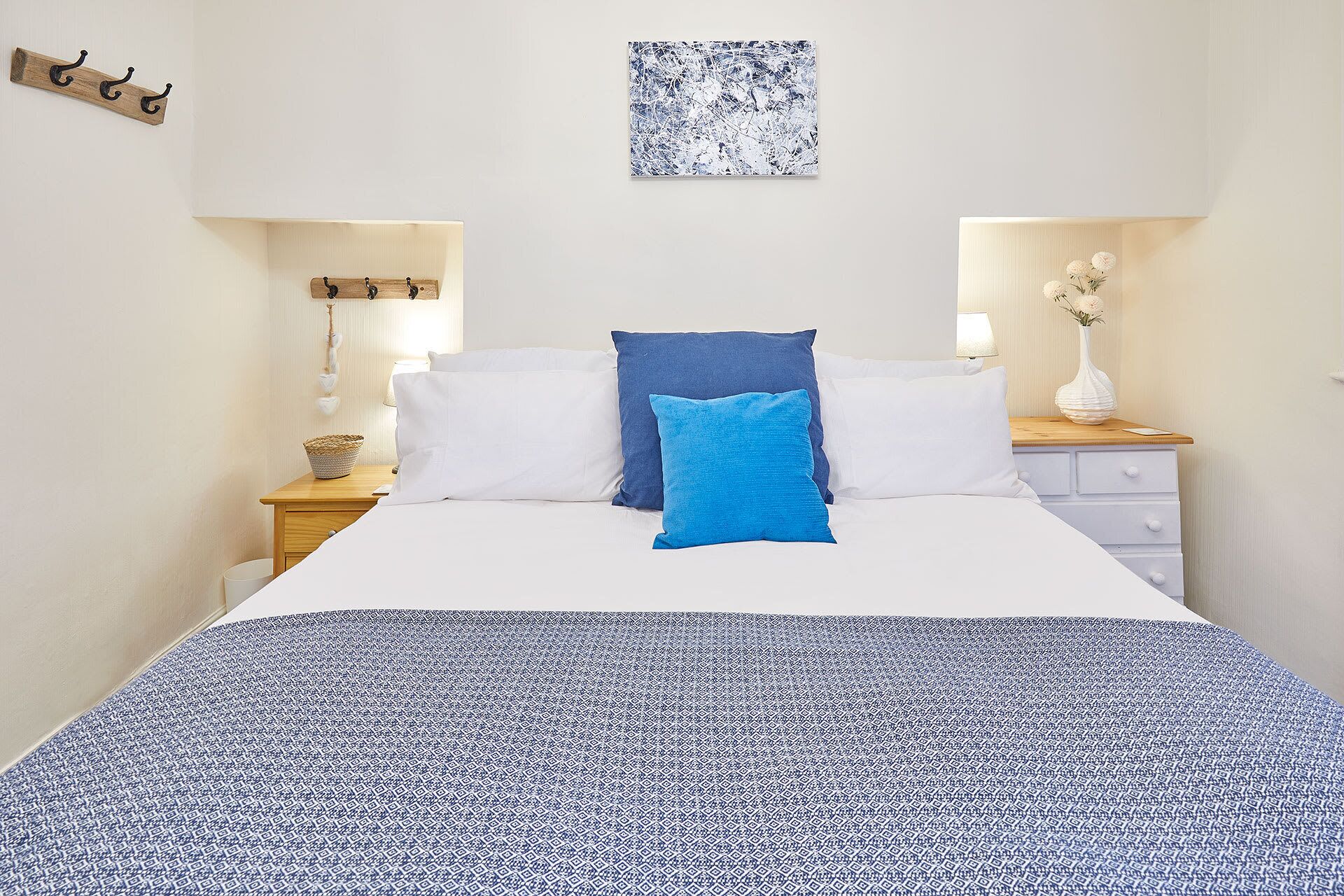 A bed with white and blue bedding, a blue pillow, and a patterned throw. There are two bedside tables with lamps and a vase of flowers.