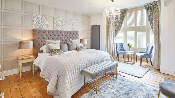 Une chambre à coucher avec une tête de lit capitonnée, un grand lit avec de nombreux oreillers, une table de chevet avec une lampe, un banc et une petite table ronde avec des chaises.