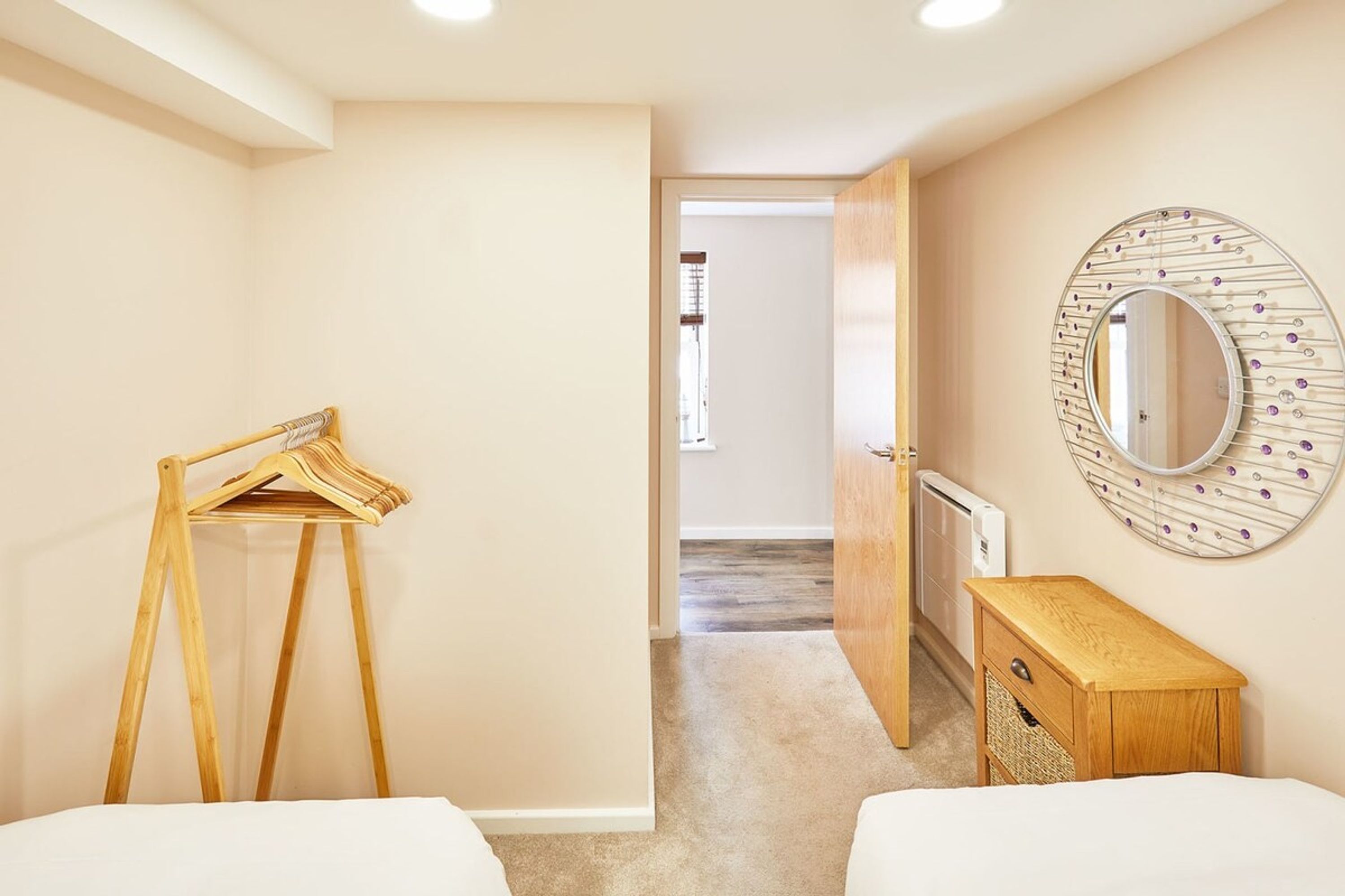 Hotellihuoneessa on puinen henkari, yöpöytä, jossa on kori, ja pyöreä peili seinällä.