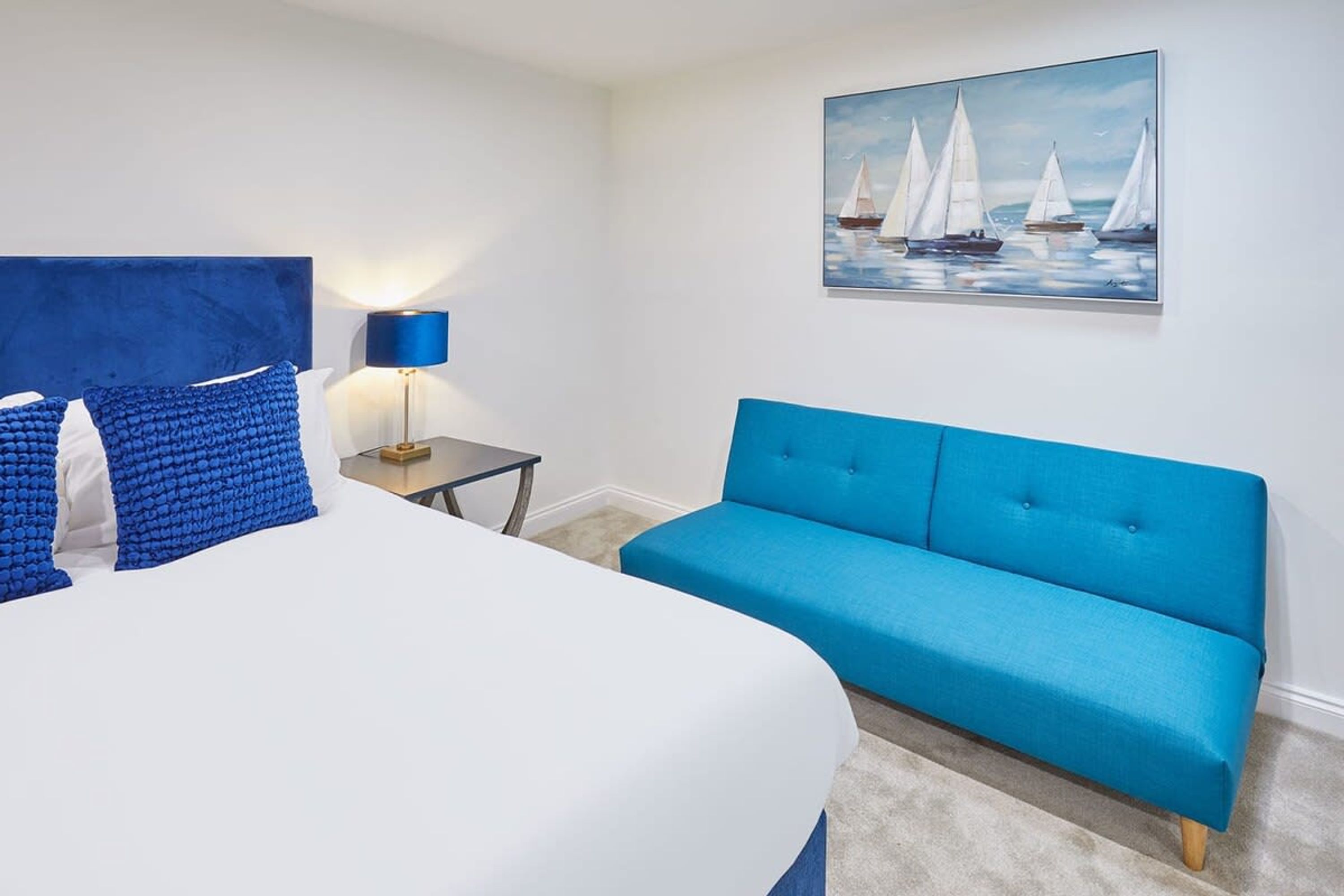 Ein Schlafzimmer mit einem großen Bett, einem blauen Sofa, einem kleinen Tisch mit Lampe und einem Segelbootgemälde an der Wand.