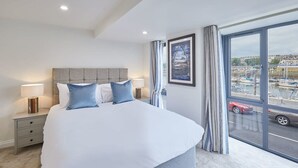 Ett modernt hotellrum med en stor säng, ett nattduksbord, en lampa och ett stort fönster med utsikt över en småbåtshamn.