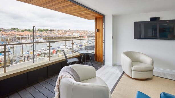 En balkong med utsikt över en småbåtshamn och stadens silhuett, inredd med en soffa, stolar och ett bord.