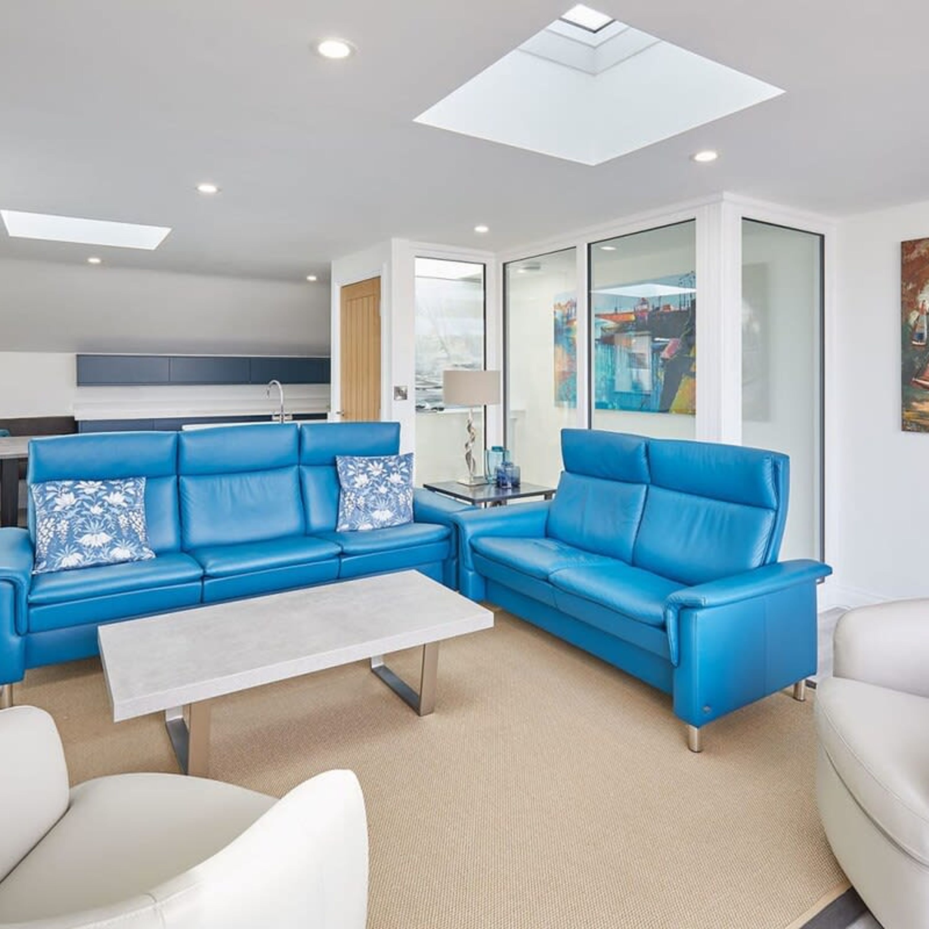 A modern living room with blue leather sofas, a white coffee table, and a glass door leading to a kitchen.