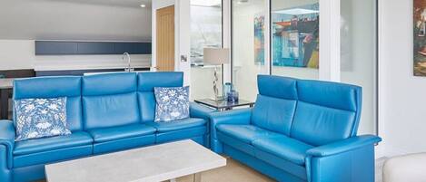 A modern living room with blue leather sofas, a white coffee table, and a glass door leading to a kitchen.