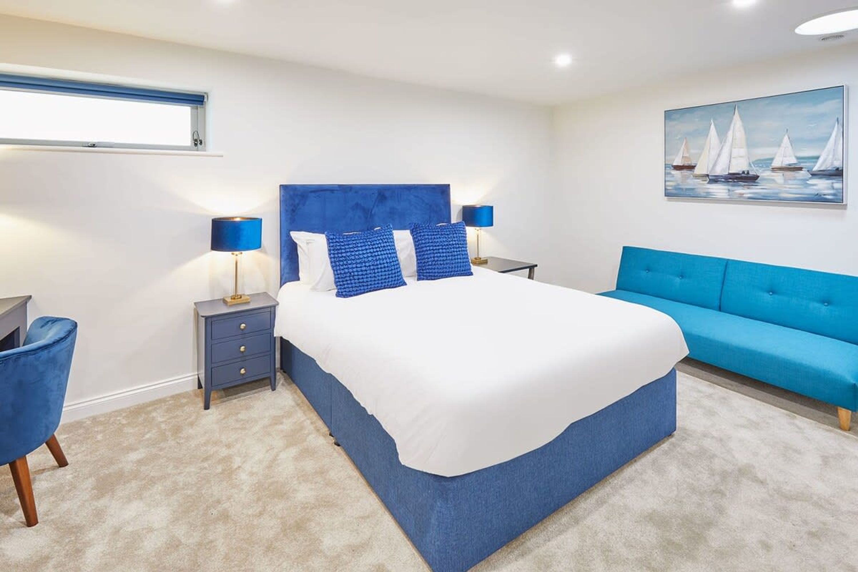A bedroom with a large bed, two bedside tables, a blue sofa, and a painting of sailboats on the wall.