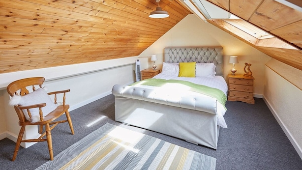 A bedroom with a sloped ceiling, a bed with a tufted headboard, a wooden chair, and a bedside table with a lamp. - Host Stay Ocean Wave (Scarborough)