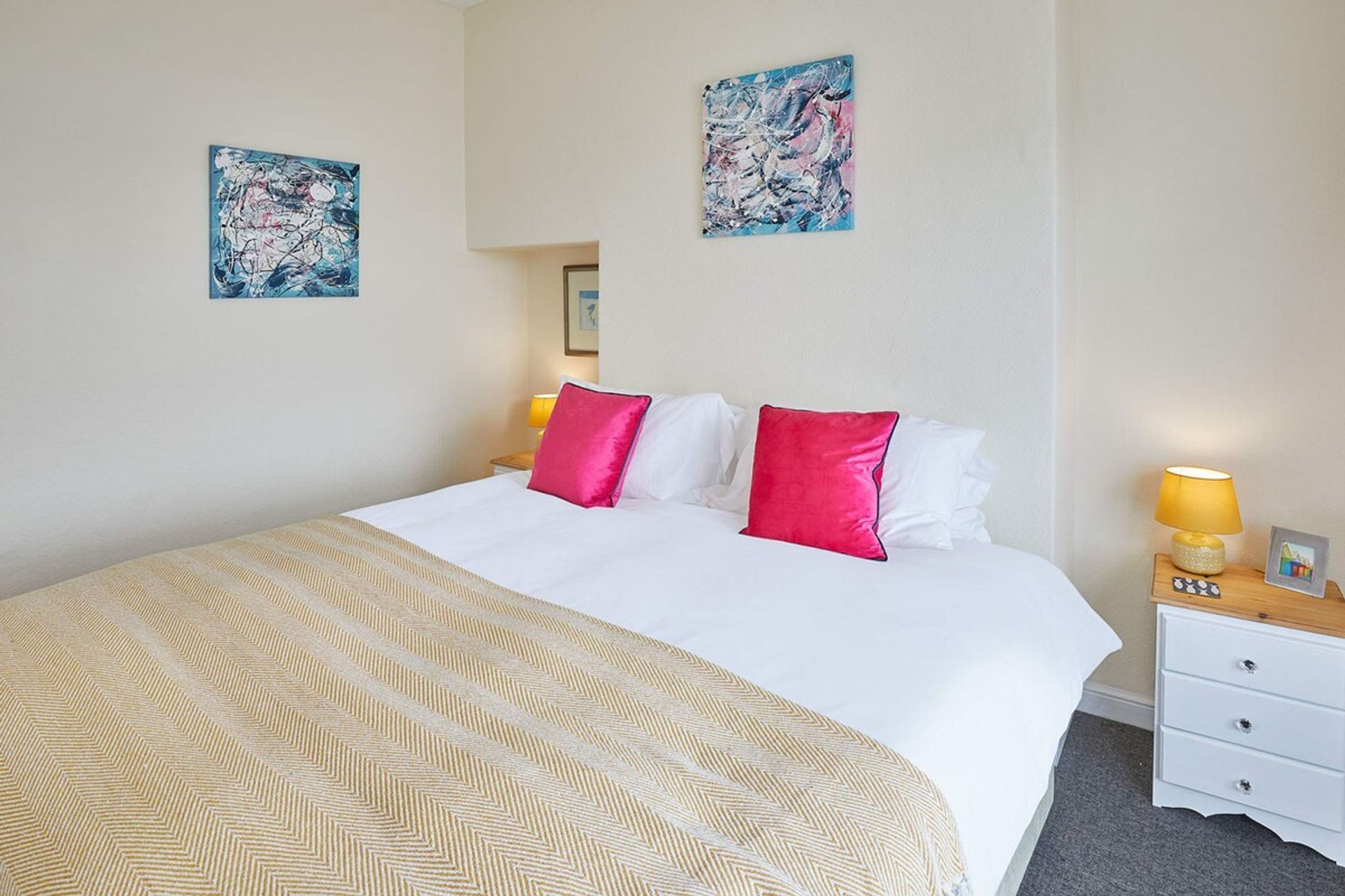 Habitación de hotel con una cama grande, dos almohadas, una mesita de noche con lámpara y dos cuadros abstractos en las paredes.