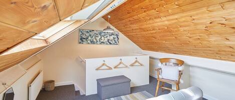 Una habitación pequeña, con paneles de madera, techo inclinado, cama, una pequeña banca, una silla y un aparador blanco con perchas.