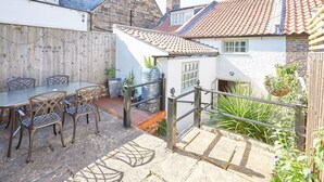 En terrasse med et bord og stole, et lille haveområde med planter, og en stentrappe der fører op til et hus med et tegltag.