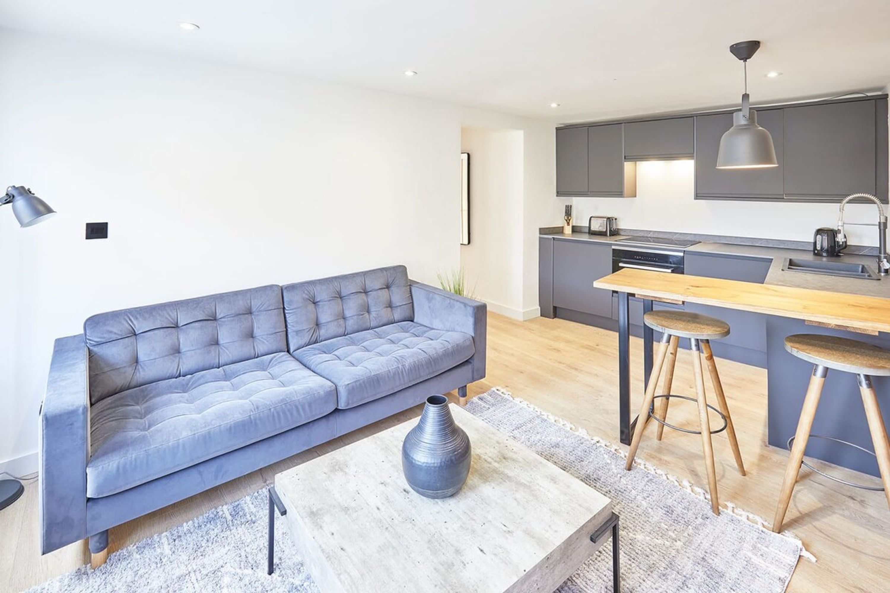 En moderne stue med en grå sofa, et træbord og et køkkenområde med en grå bordplade og barstole.