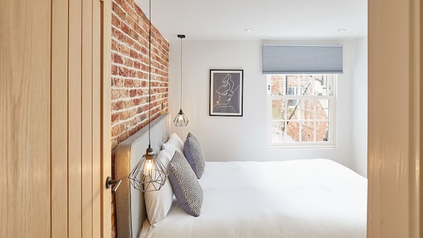 A bedroom with a brick wall, a bed with white bedding, a framed picture, and a window with blinds. - Host Stay Modern Industrial Golden Lion Apartments (Whitby)