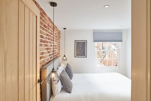 Une chambre avec un mur en briques, un lit avec une literie blanche, un tableau encadré et une fenêtre avec des stores.