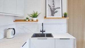 A modern kitchen with white cabinetry, a black faucet, and a marble countertop. There are two potted plants on a wooden shelf above the sink. - Host Stay Simply Scandi Golden Lion Apartments (Whitby)