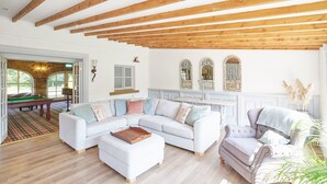 Een ruime woonkamer met een witte hoekbank, een grijze fauteuil en een houten plafond.