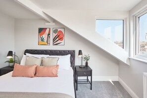 Un dormitorio con cama, dos obras de arte en la pared, una mesita de noche, una lámpara y una ventana con vistas.