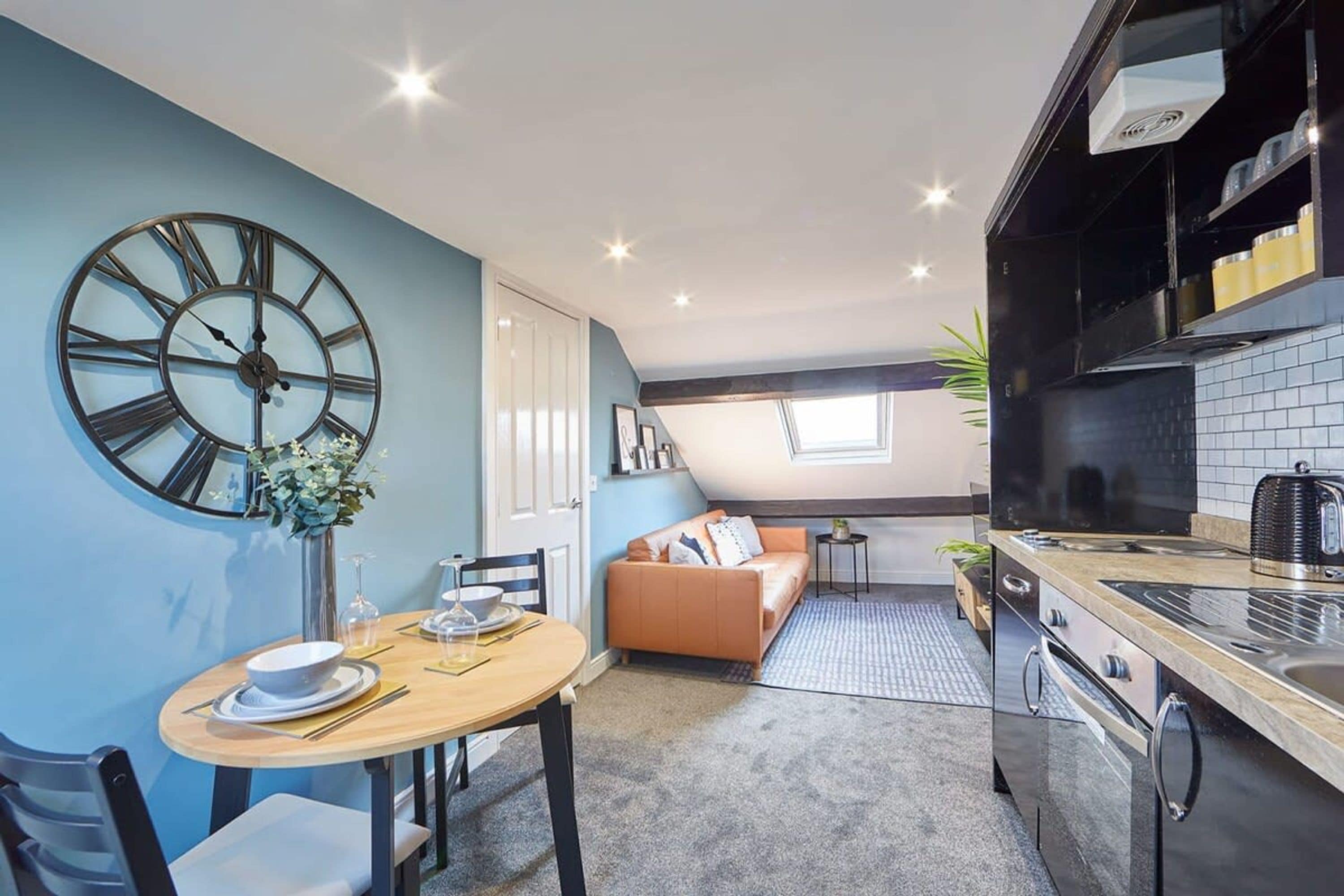 A modern kitchen with a large clock, a dining area with a table set for two, and a cozy living space with a sofa and a small side table.