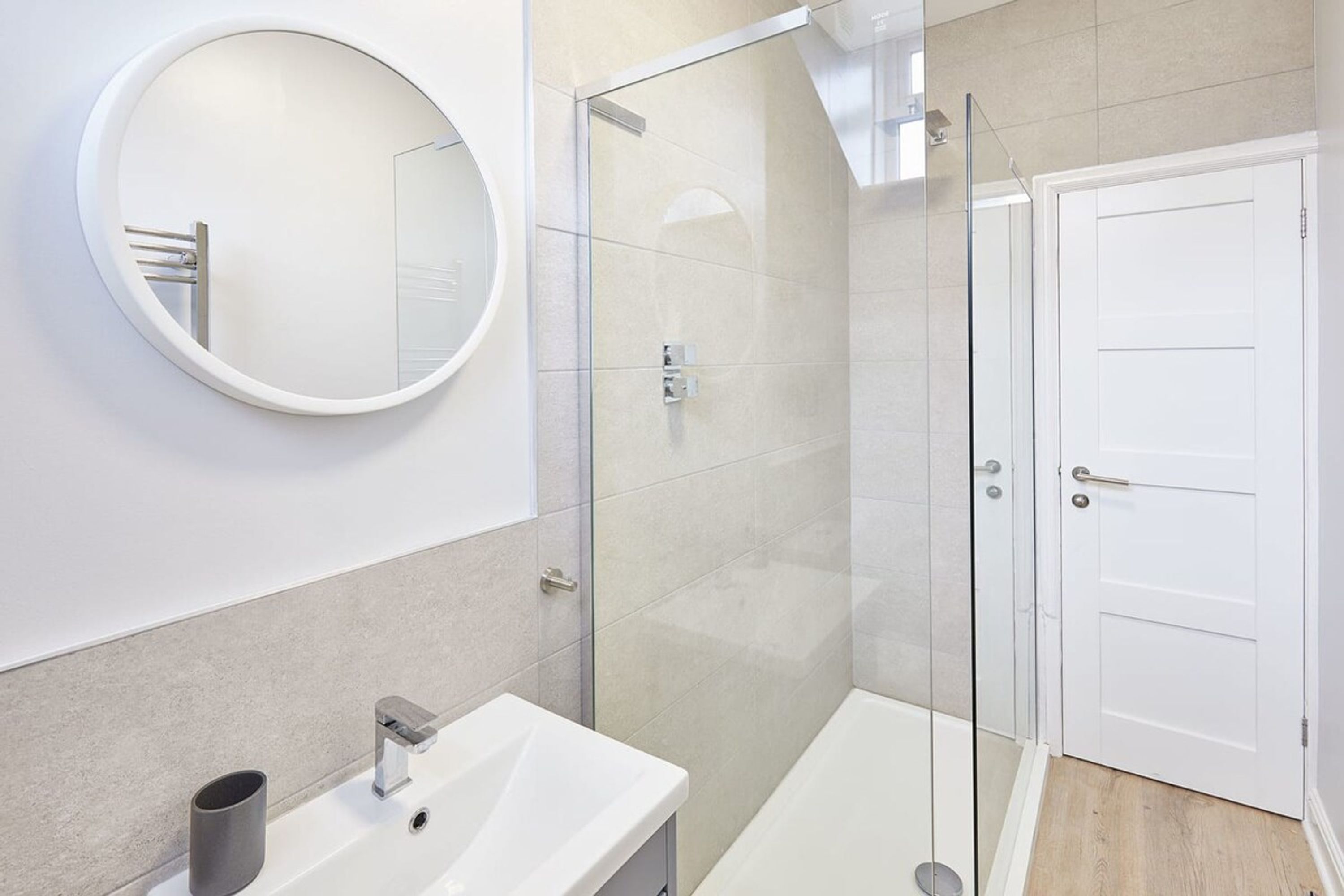 A modern bathroom with a round mirror, a white sink, a glass-enclosed shower, and a white door.