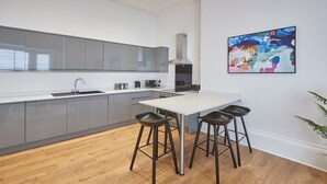 A modern kitchen with grey cabinets, a white countertop, and a stainless steel range hood. There are three black bar stools at the island. - Host Stay Sandy Cove (Saltburn-by-the-Sea)