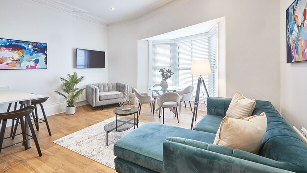 A modern living room with a teal sofa, a grey armchair, a glass coffee table, and a dining area with a white table and black chairs. - Host Stay Sandy Cove (Saltburn-by-the-Sea)