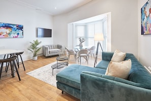 Ein modernes Wohnzimmer mit einem türkisfarbenen Sofa, einem grauen Sessel, einem Glastisch und einem Essbereich mit weißem Tisch und schwarzen Stühlen.