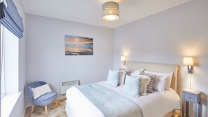 A bedroom with a large bed, a chair, a bedside table, and a wall-mounted lamp. - Host Stay Starboard Bow (Whitby)