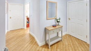 A hallway with wooden flooring, white doors, a light-colored console table with a mirror, and a vase with flowers. - Host Stay Starboard Bow (Whitby)