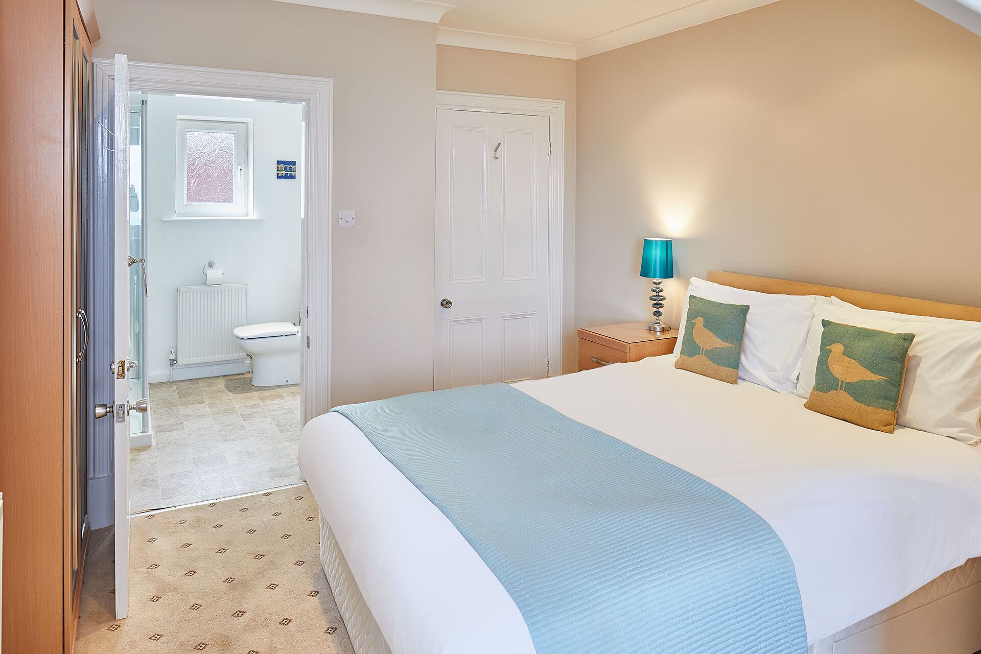 A hotel room with a bed, a nightstand with a lamp, a bathroom with a toilet and a radiator, and a door leading to another room.