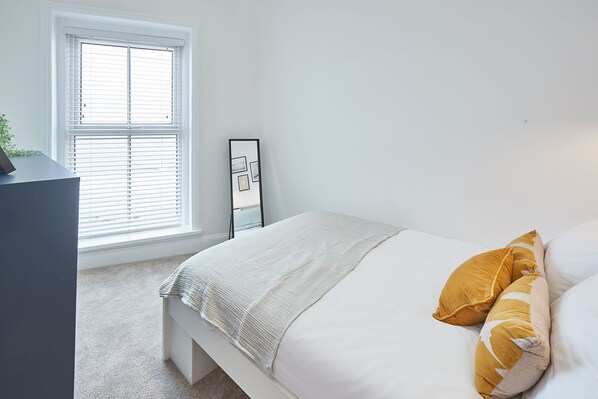 A bedroom with a bed, a bedside table, a window with blinds, and a wall-mounted mirror. - Host Stay Anchors Away (Saltburn-by-the-Sea)