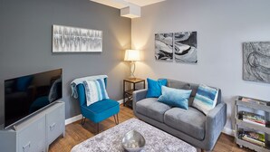 Un salon moderne avec un canapé gris, un fauteuil bleu, un téléviseur à écran plat et des œuvres d’art accrochées aux murs.