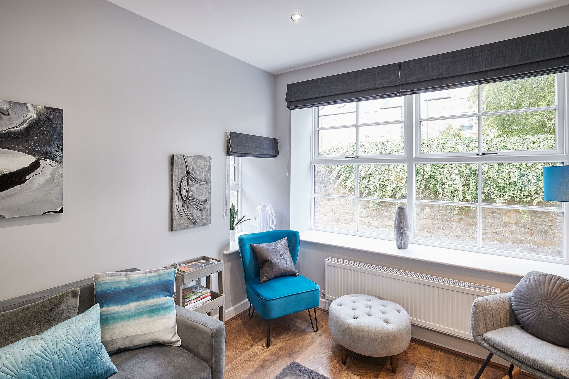 A modern living room with a blue armchair, grey sofa, and ottoman. There is a large window with a view of greenery outside.