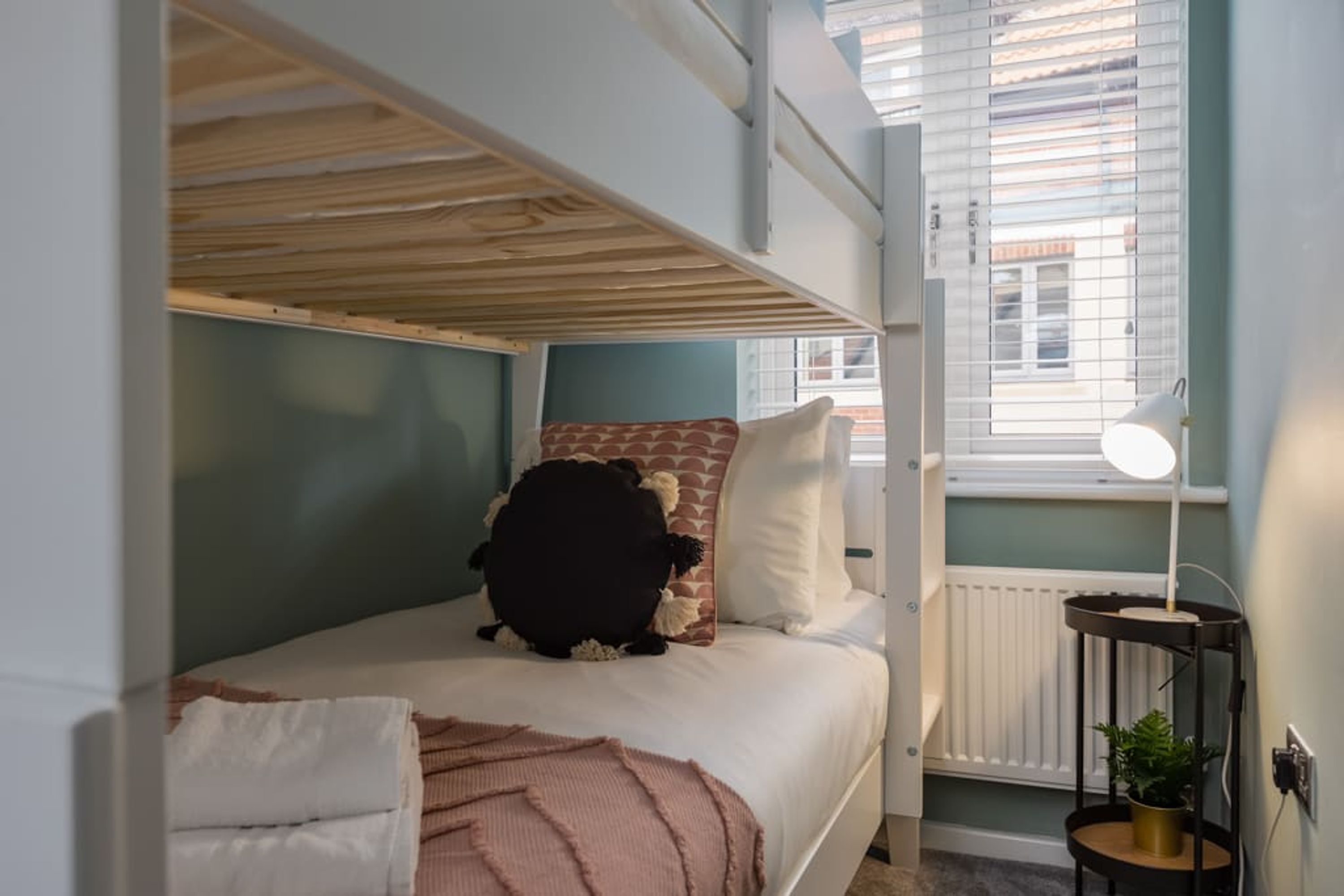 Un lit superposé avec un matelas et des oreillers blancs, une petite table de chevet avec une lampe et une fenêtre avec des stores.