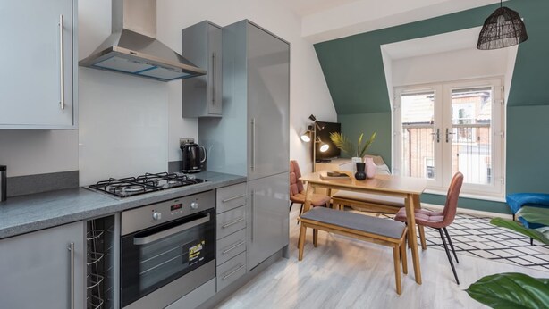 A modern kitchen with a gas stove, oven, and built-in cabinets. There is a dining area with a wooden table and chairs, and a window with a view of buildings.