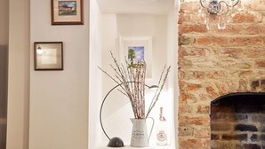 A room with a brick fireplace, framed pictures on the wall, a vase with branches, and a candle. - Host Stay Willow Cottage (York)